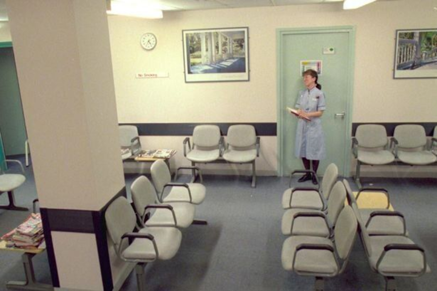 A mostly empty waiting room with rows of gray chairs, a nurse in uniform standing near the door reading a file, wall art, a clock, and magazines on small tables.