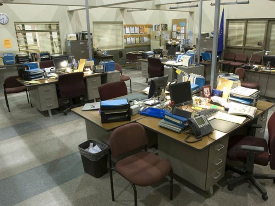 A large, open-plan office with several desks arranged in clusters, covered with paperwork, office supplies, computers, and phones. Empty chairs are placed at each desk, and the room appears tidy but unoccupied.