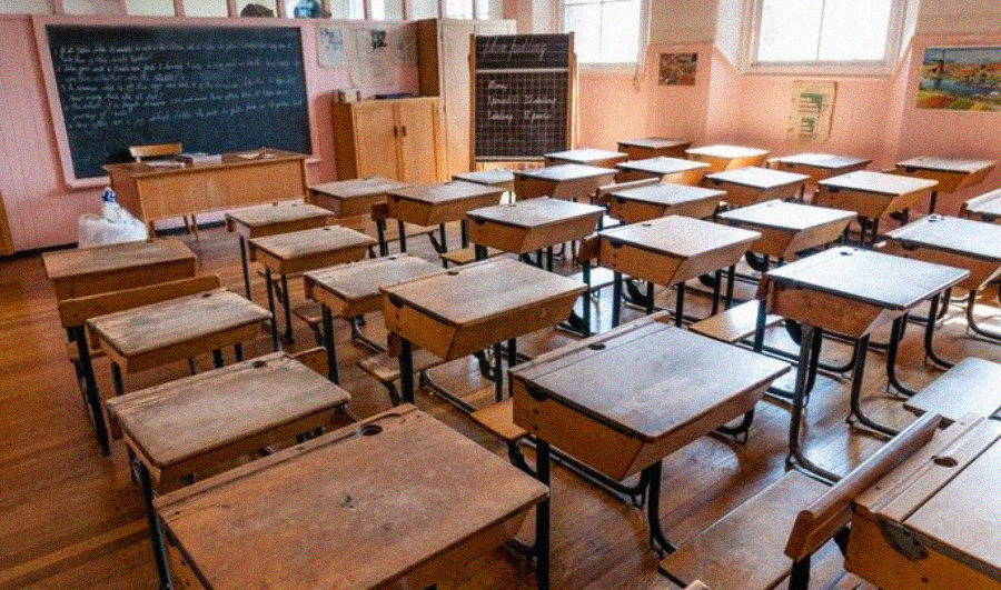 An empty classroom with rows of wooden desks and chairs, a blackboard with writing on it, cabinets, and large windows letting in sunlight. The room appears tidy but unoccupied.