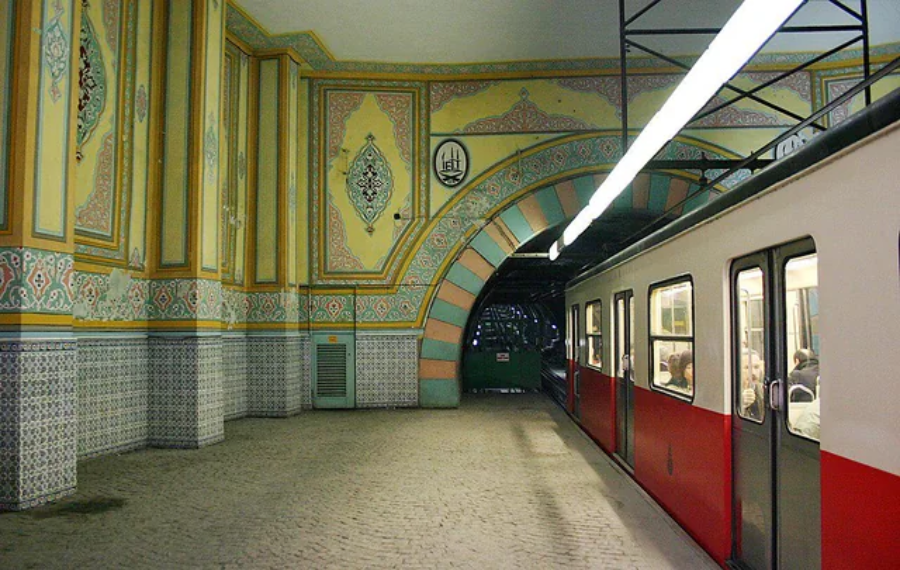 A red and white subway train is stopped at an ornate underground station with patterned tile walls, archways, and intricate yellow, green, and orange decorative designs. Passengers are visible through the windows.