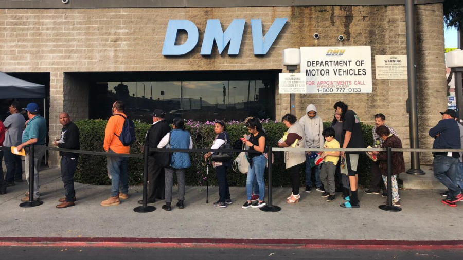 A diverse group of people stand in a long line outside a DMV building, waiting for service. The DMV sign is visible above the entrance, and a large notice board is displayed next to the doors.