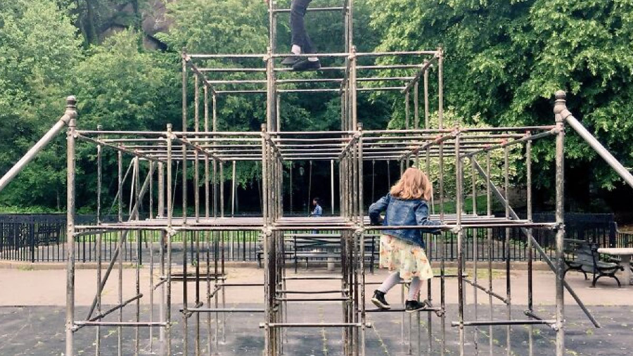 A young girl climbs on a large metal jungle gym in a park. Trees and benches are visible in the background. The scene appears calm and playful.