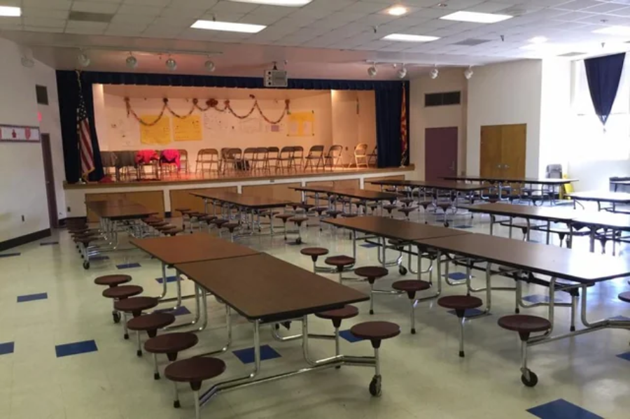 A school cafeteria with rows of long brown tables and round stools, facing a stage decorated with garlands and chairs. The room is empty and well-lit with overhead lights.