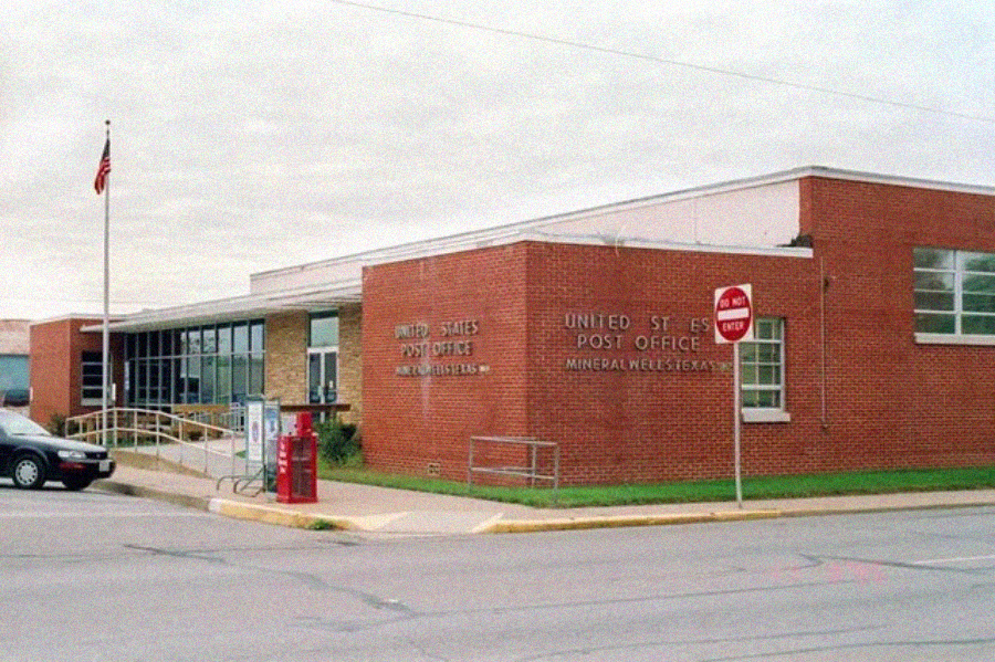 A one-story red brick United States Post Office building sits on a street corner, with a ramp, American flag, and stop sign visible in front. The sky is overcast and a car is parked nearby.