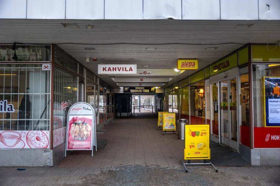 A covered walkway between two shops; on the left is a café with “KAHVILA” sign, and on the right is a grocery store “alepa.” Promotional signs and a menu board stand outside the entrances.