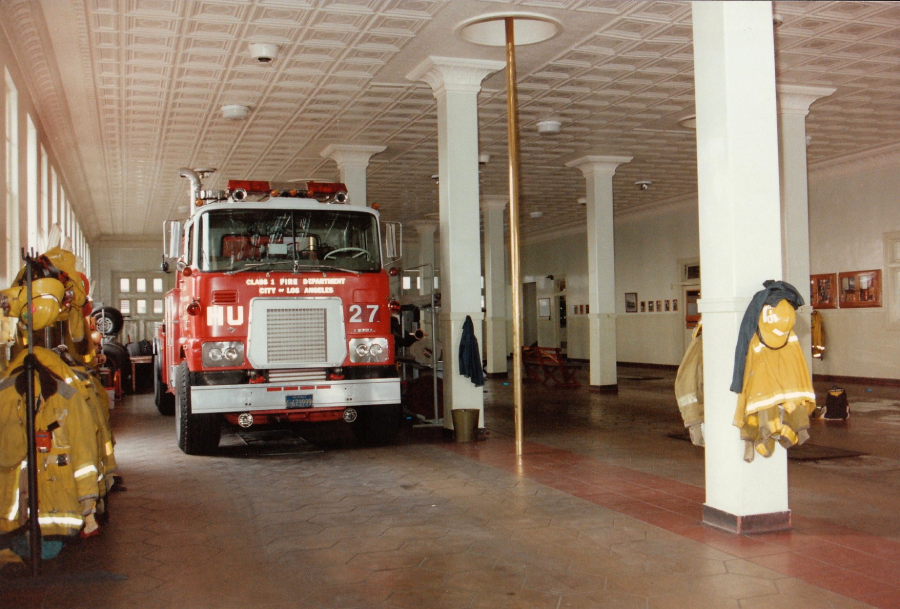 A red fire truck is parked inside a fire station with tiled floors and high ceilings. Firefighting gear hangs on the wall to the left, and a brass fire pole is in the center of the room.