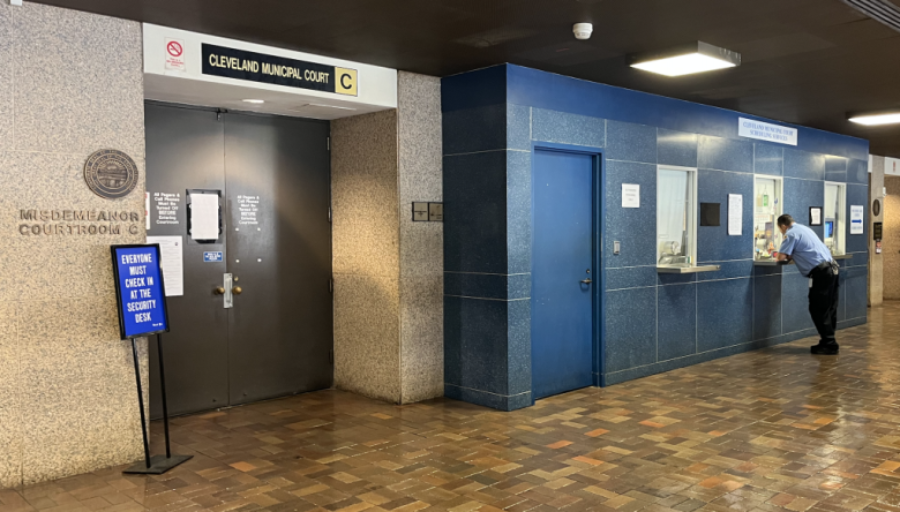 A hallway in Cleveland Municipal Court featuring a blue information desk where a man speaks to a staff member, a sign for Courtroom C, and a security checkpoint sign near double doors.