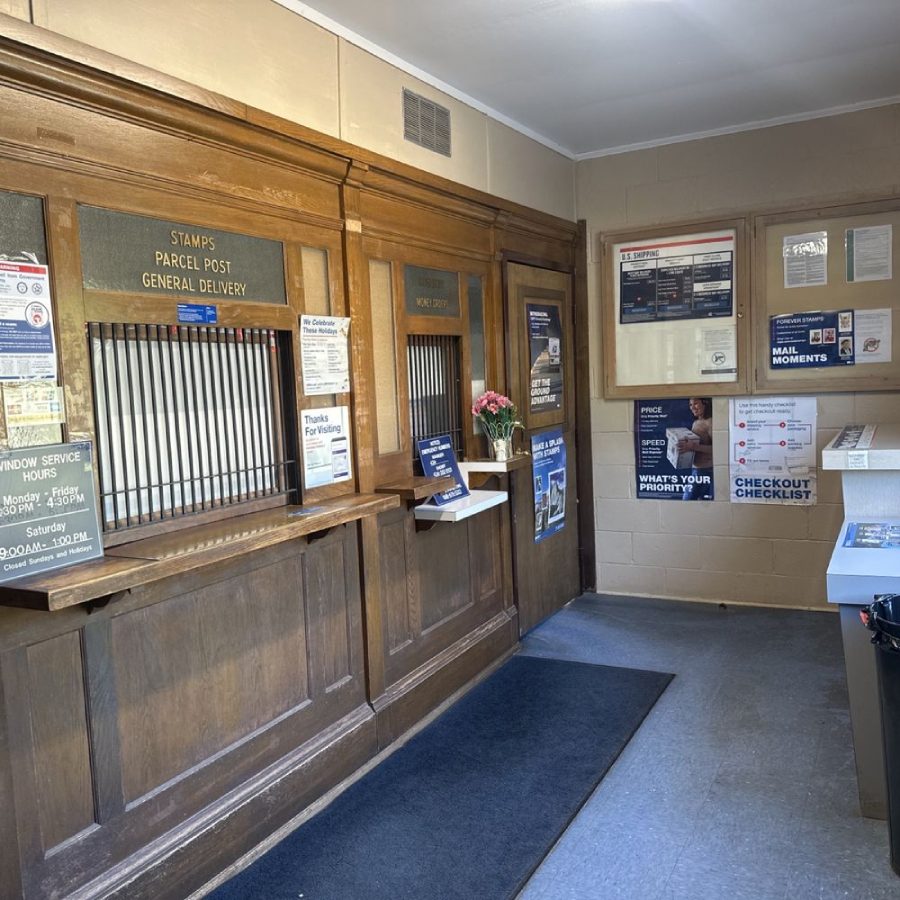 A small post office lobby with wooden service windows, informational posters on the walls, a counter with forms, and a vase of flowers on a shelf beside a transaction window.