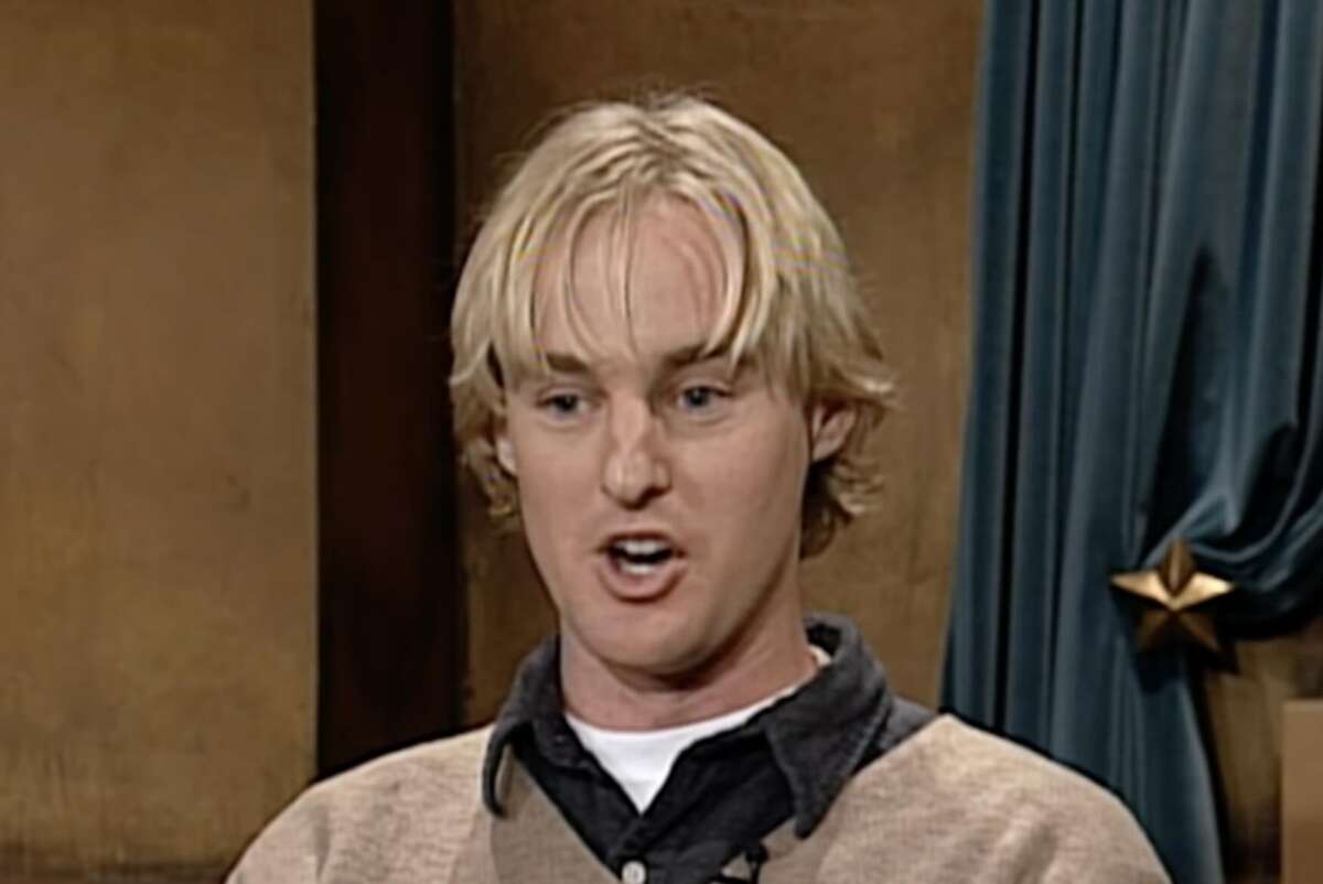 A man with blonde hair wearing a collared shirt and sweater sits indoors, speaking. There is a tan wall and a blue curtain with a gold star decoration behind him.