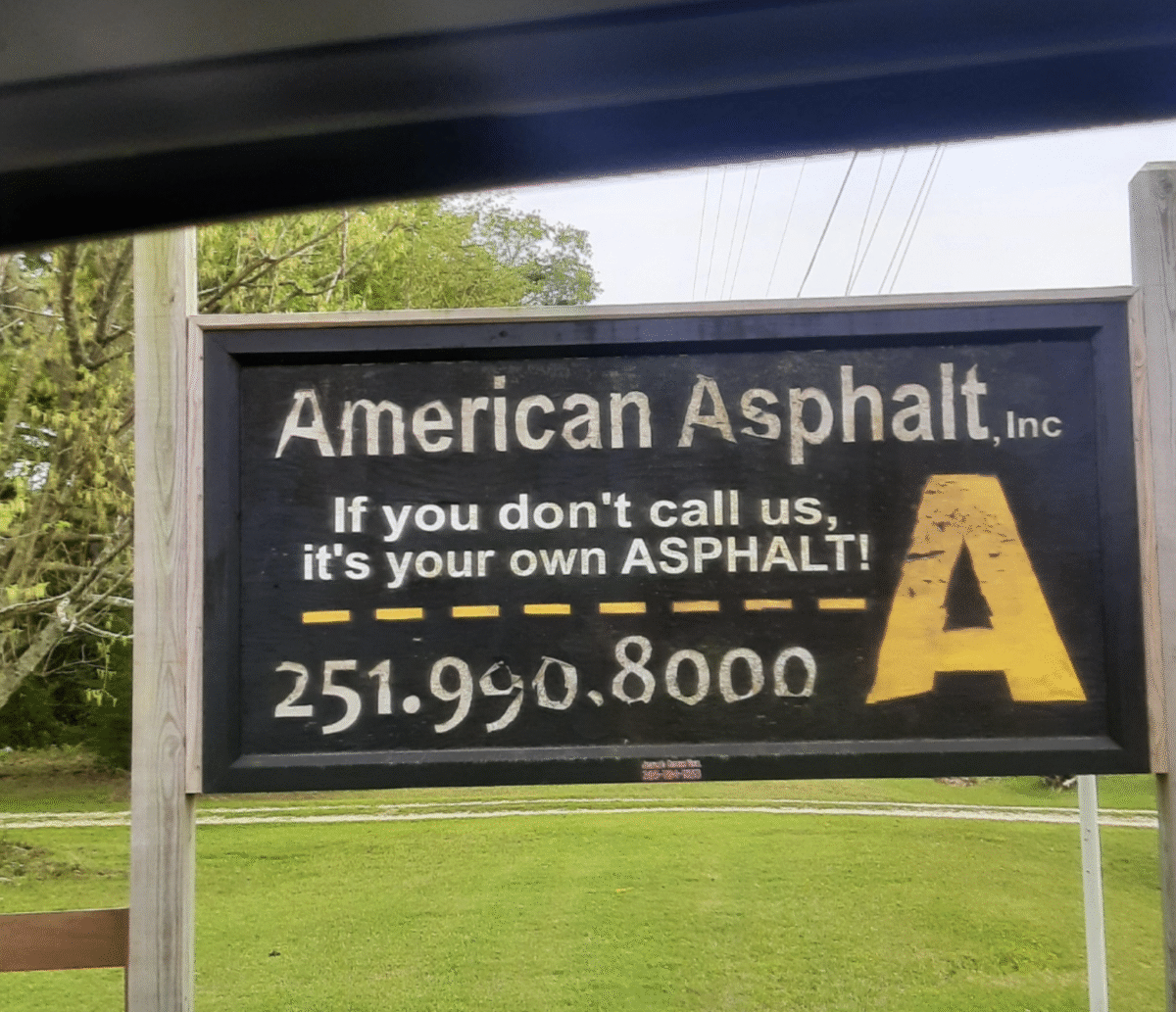 A roadside sign for "American Asphalt, Inc" reads: "If you don’t call us, it’s your own ASPHALT!" with the phone number 251.990.8000 and a large yellow letter "A" on the right. Trees and grass are in the background.