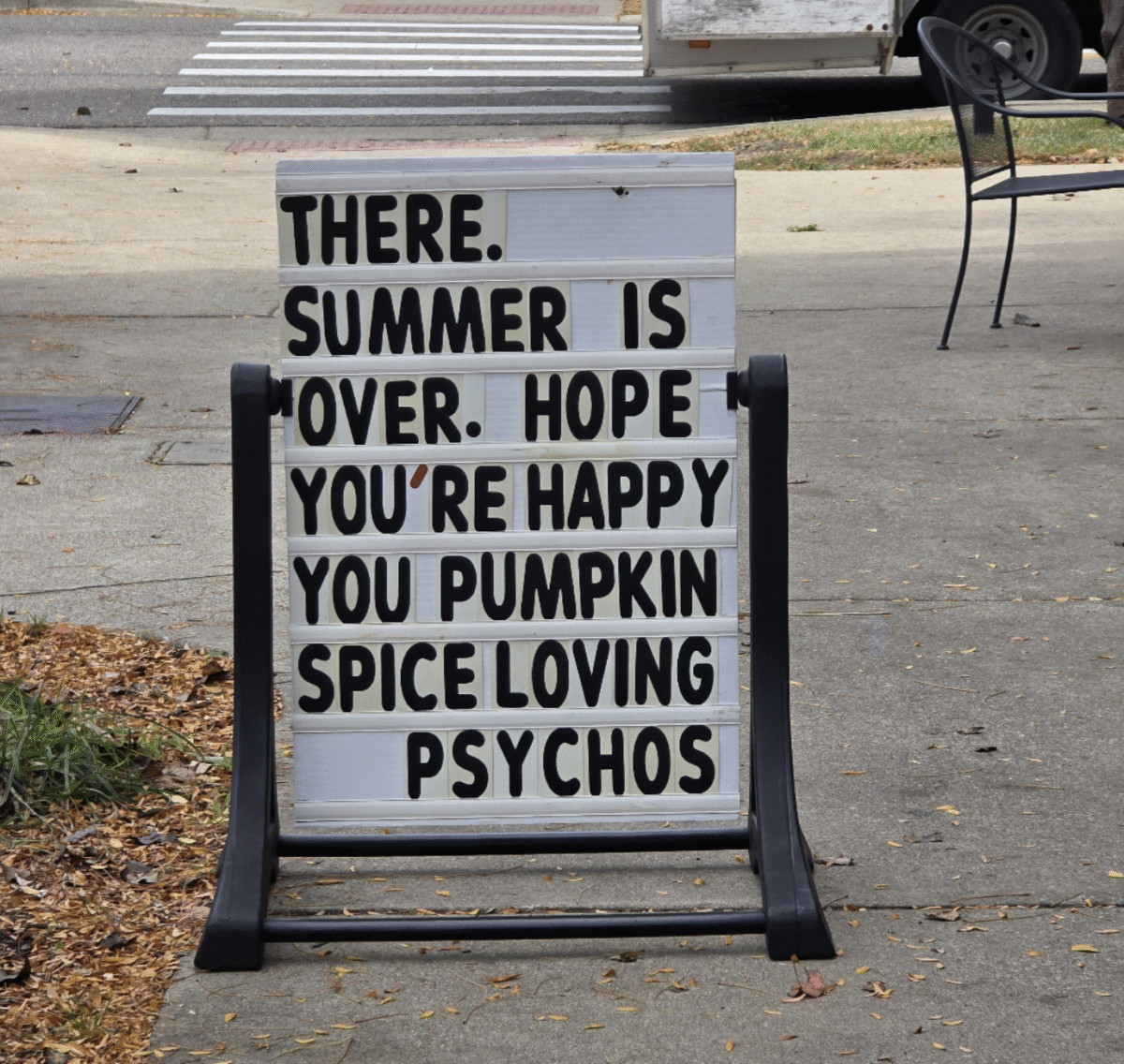 A sidewalk sign reads: "THERE. SUMMER IS OVER. HOPE YOU'RE HAPPY YOU PUMPKIN SPICE LOVING PSYCHOS." The sign is placed near a street with fallen leaves nearby.