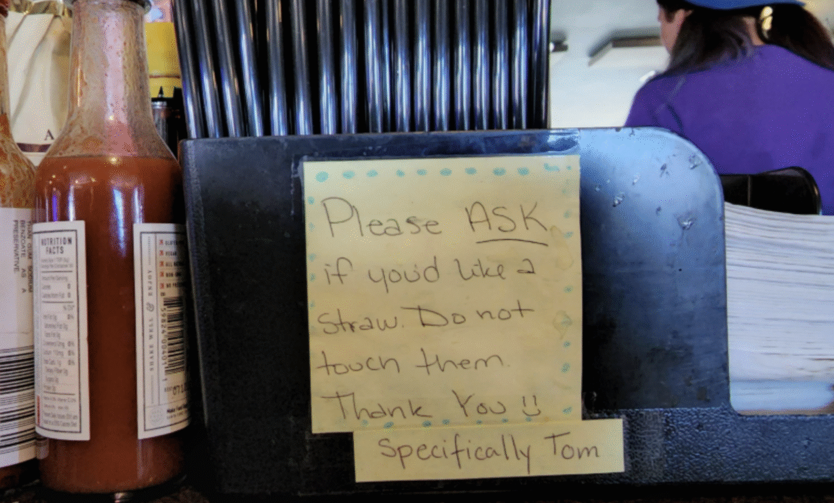A handwritten note on a napkin holder reads: "Please ASK if you'd like a straw. Do not touch them. Thank you!! Specifically Tom." Condiment bottles and straws are visible nearby.
