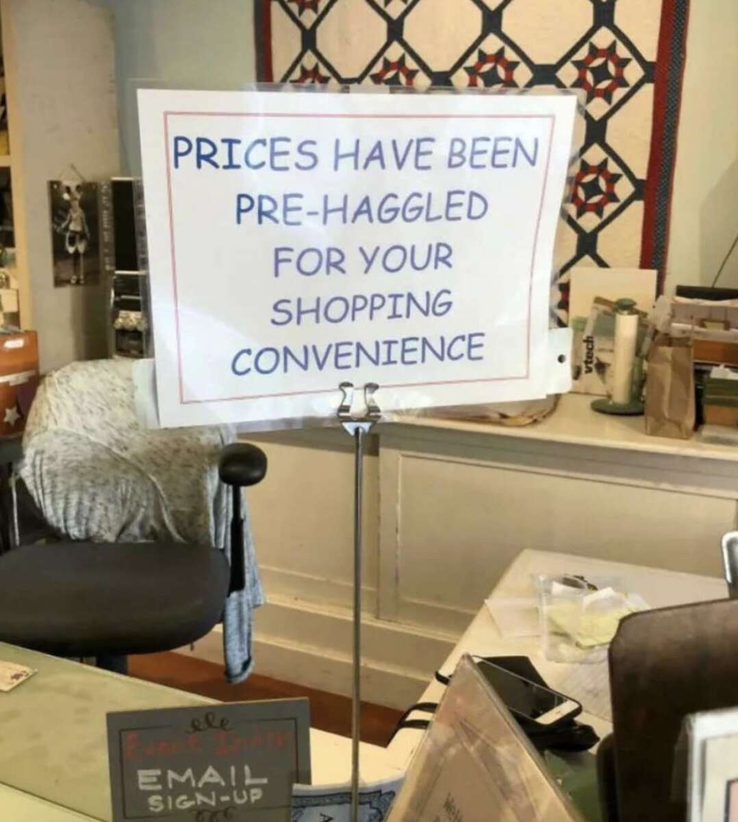 A sign on a shop counter reads, "PRICES HAVE BEEN PRE-HAGGLED FOR YOUR SHOPPING CONVENIENCE." The counter has office supplies, papers, and a chair behind it.
