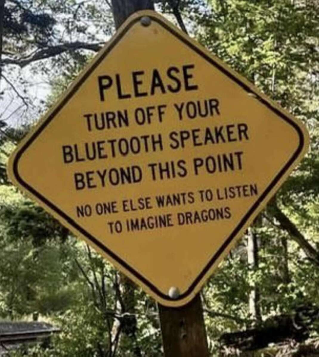 A yellow diamond-shaped sign in a wooded area reads: "PLEASE TURN OFF YOUR BLUETOOTH SPEAKER BEYOND THIS POINT. NO ONE ELSE WANTS TO LISTEN TO IMAGINE DRAGONS.