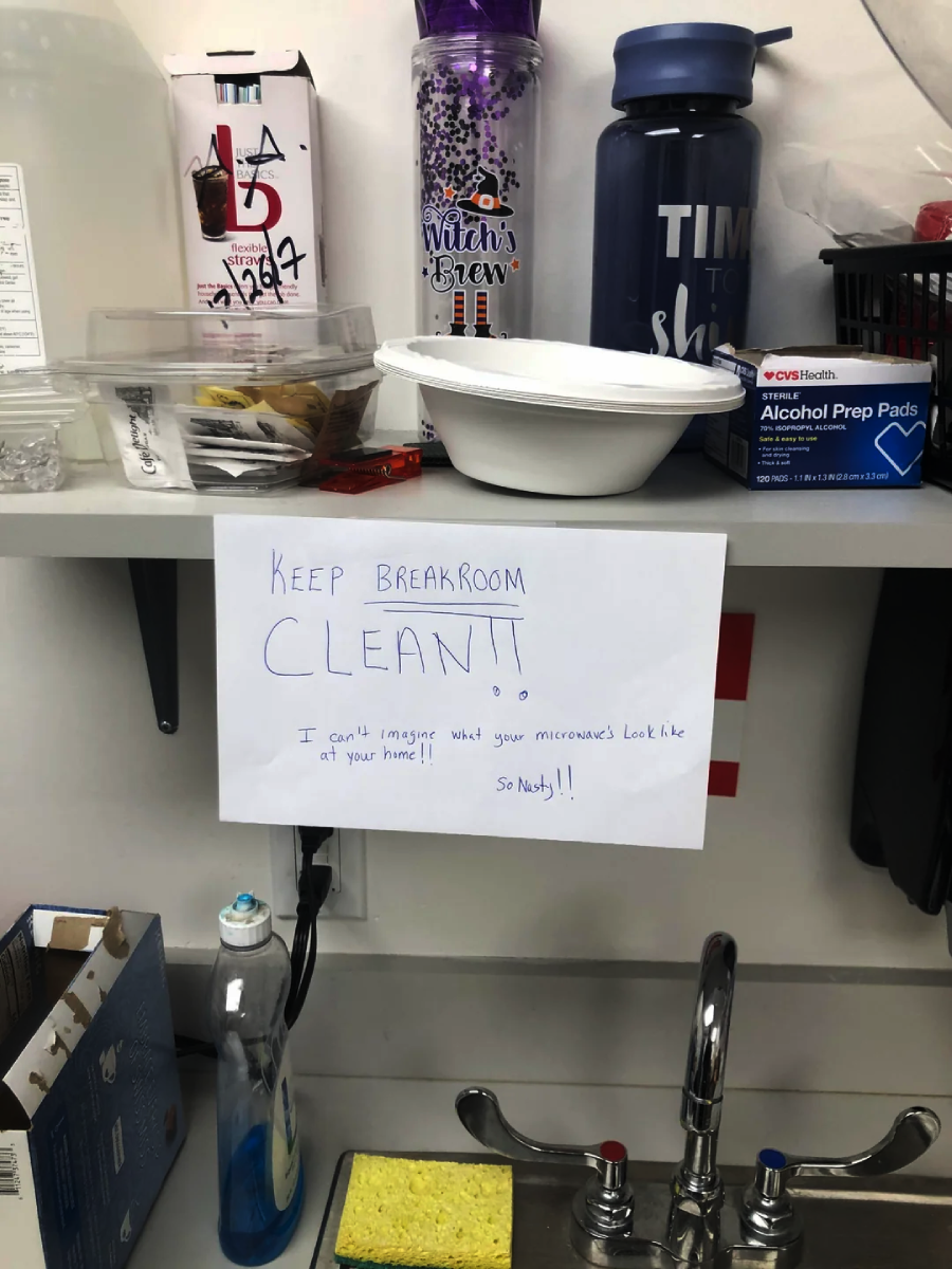 A shelf above a sink holds snacks, cups, and bottles. A handwritten sign says, “KEEP BREAKROOM CLEAN!! I can’t imagine what your microwave looks like at your home!! So nasty!!” A box of alcohol prep pads is also visible.