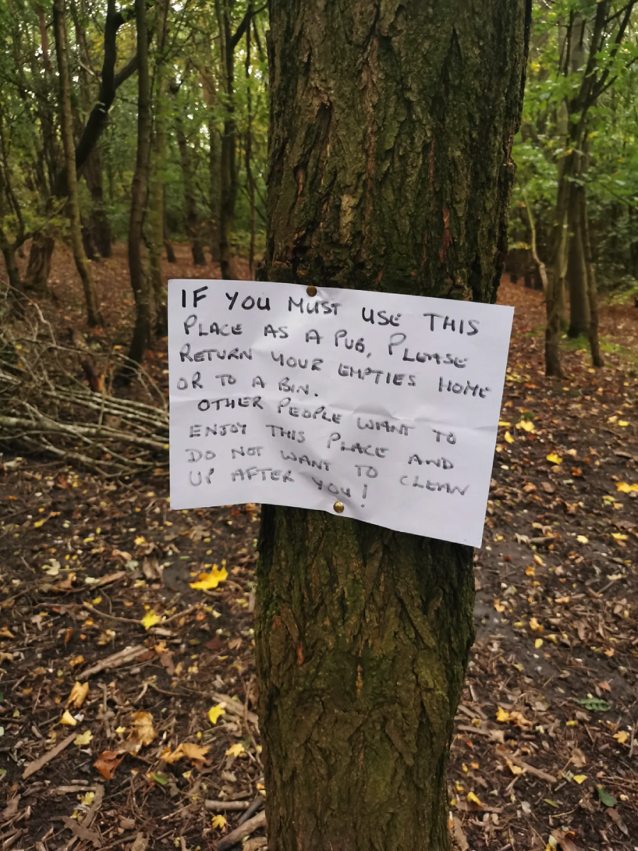 A handwritten sign is pinned to a tree in a forest. The sign asks people to clean up after themselves and not leave litter, reminding them that others want to enjoy the area too. The forest floor is covered with leaves.