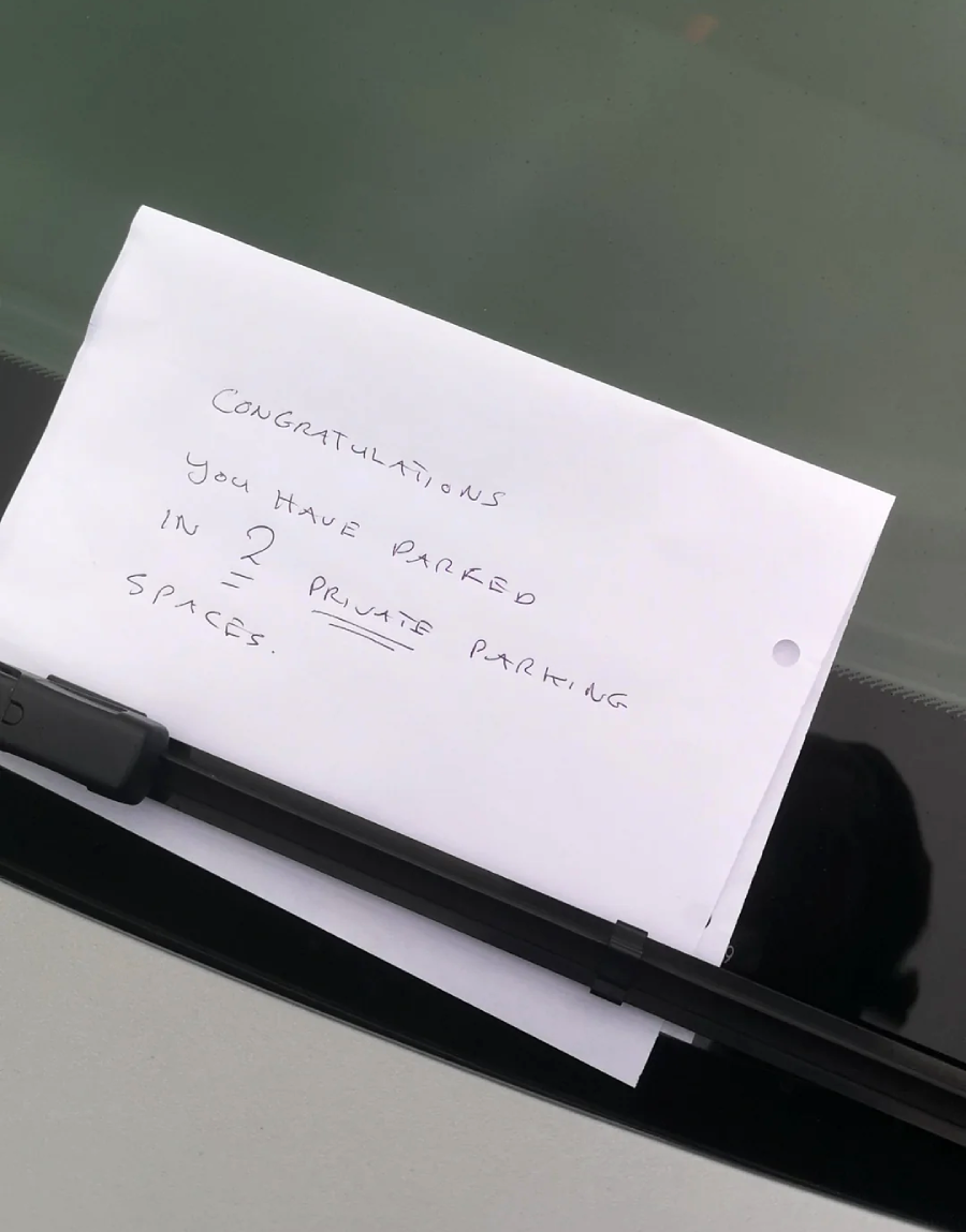 A handwritten note under a car windshield wiper reads: "CONGRATULATIONS YOU HAVE PARKED IN 2 PRIVATE PARKING SPACES.