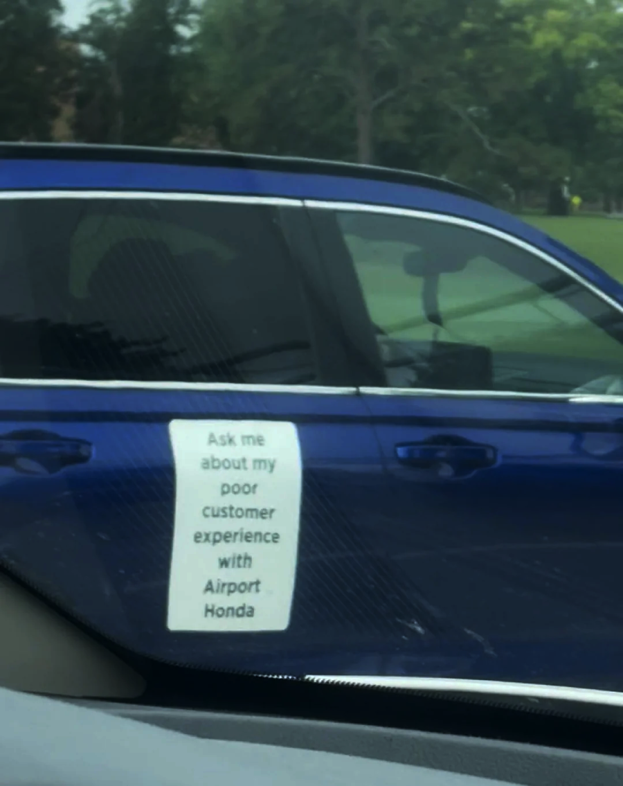 A blue car with a large white sign on the driver's door that reads, "Ask me about my poor customer experience with Airport Honda." Trees and grass are visible in the background.