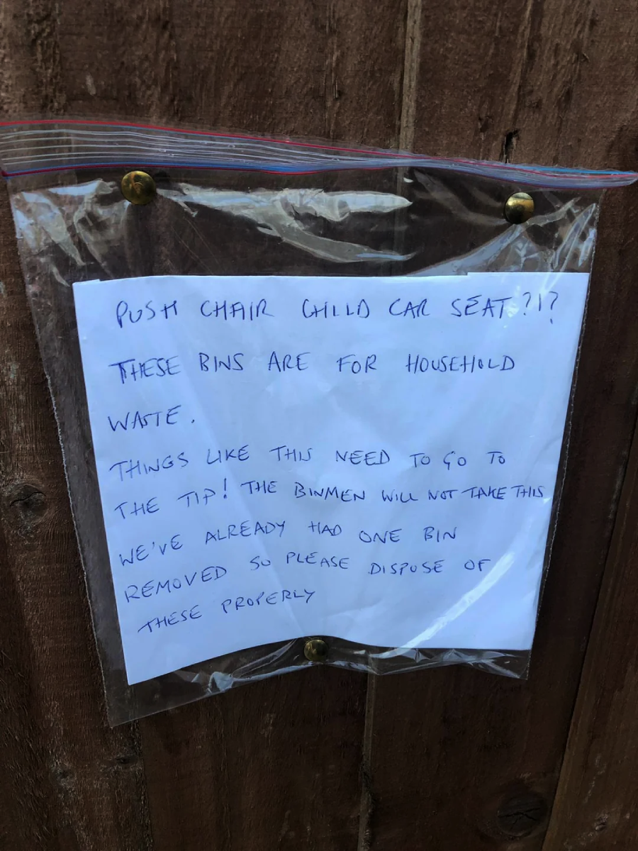 A handwritten note in a plastic sleeve is pinned to a wooden fence. The note asks people not to put pushchairs, child car seats, or similar items in household bins and requests proper disposal at the tip.