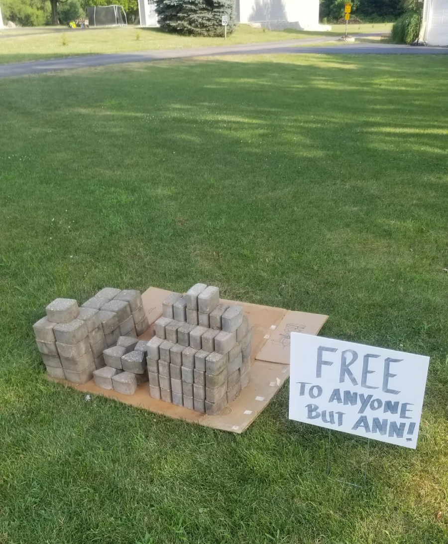 A pile of concrete pavers sits on cardboard on a lawn. A handwritten sign beside them reads, "FREE TO ANYONE BUT ANN!" A street and houses are visible in the background.