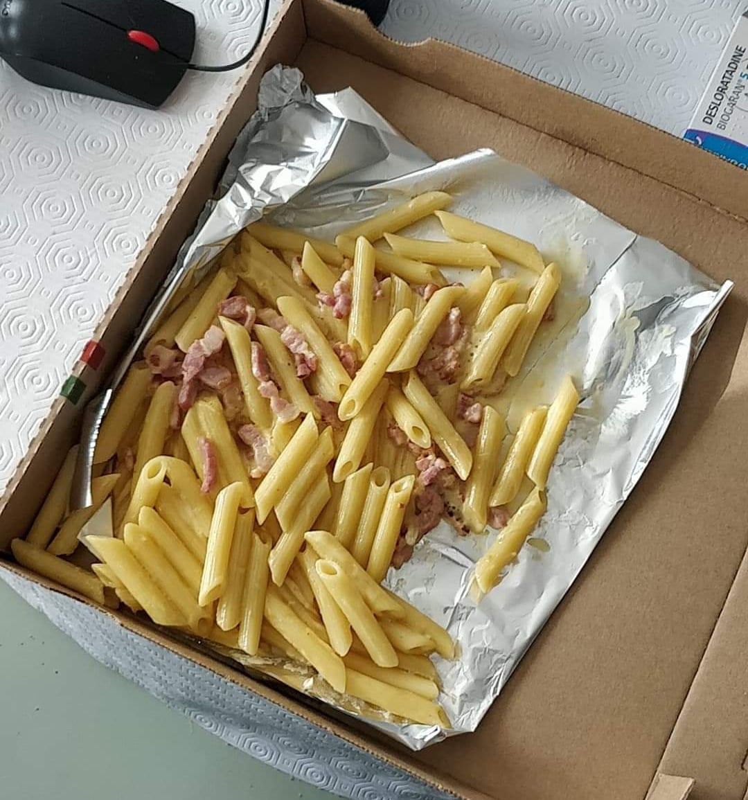 A cardboard pizza box lined with aluminum foil contains a sparse serving of penne pasta mixed with a few pieces of bacon. A fork rests on the foil inside the box. A computer mouse is visible nearby.