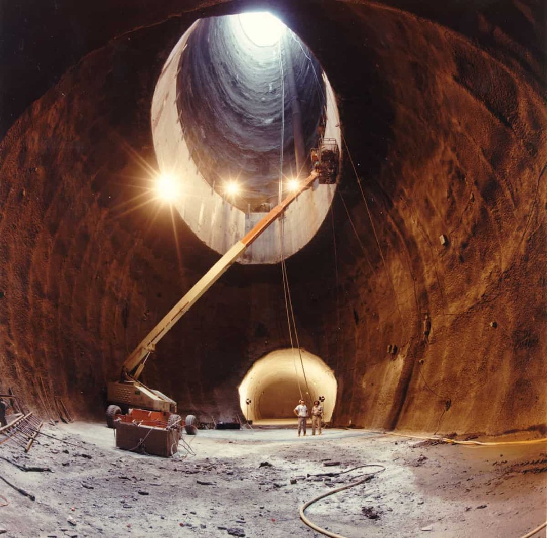 Construction workers stand at the base of a large, cylindrical underground tunnel. Bright lights illuminate the rough walls, and a crane extends upward toward a circular opening at the tunnel’s top, letting daylight in.