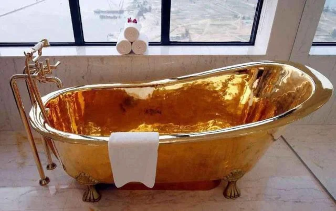 A shiny gold bathtub with claw feet sits by a large window. A white towel hangs over the side, and three rolled white towels are stacked on the windowsill, with a city view visible outside.