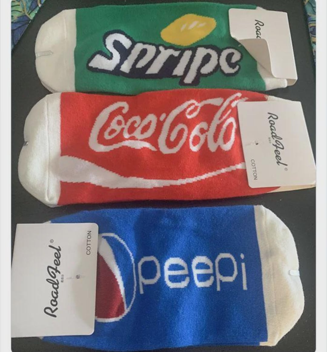 Three pairs of socks mimic soda logos: green with "Snipe" (Sprite), red with "Coco-Cole" (Coca-Cola), and blue with "peepi" (Pepsi). Each has a white tag labeled "RoadJeel.