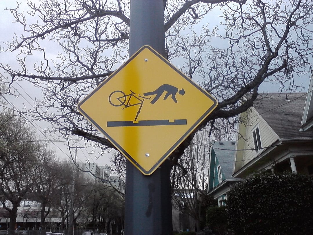 A yellow warning sign on a pole shows a stick figure falling off a bicycle after hitting a raised obstacle, with trees and houses in the background.