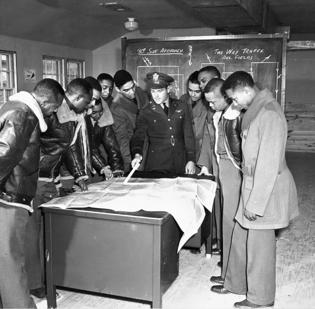 A military officer in uniform points to a map on a desk while a group of young men closely watch. They are gathered in a classroom with a chalkboard displaying aviation instructions in the background.