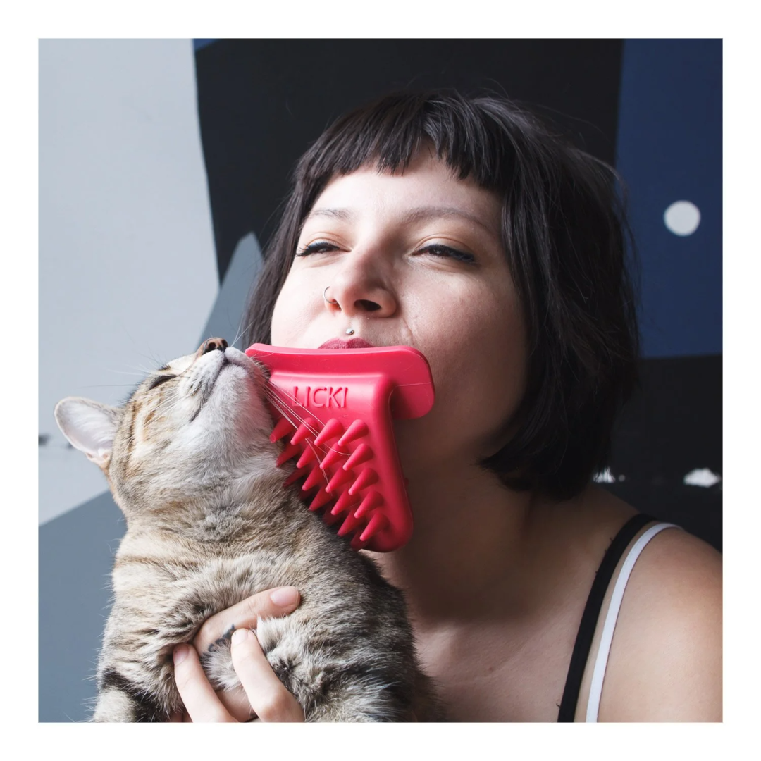 A person uses a red, brush-like device held in their mouth to “lick” a gray tabby cat, which they are holding close, mimicking grooming behavior. The cat looks content.