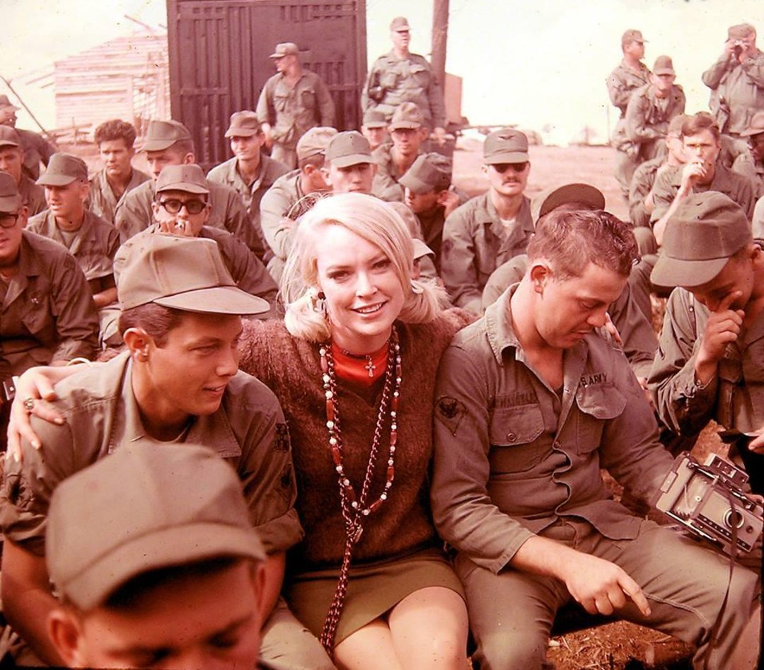 A woman with blonde hair sits smiling among a group of U.S. soldiers in uniform, many of whom are seated and looking in different directions outdoors. The mood appears relaxed and informal.