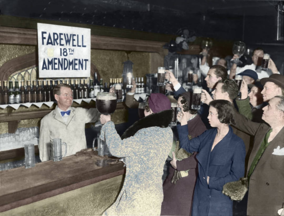 A group of people raise their glasses in celebration at a bar with a sign that reads “Farewell 18th Amendment,” marking the end of Prohibition in the United States. Bottles line the shelves behind the bartender.