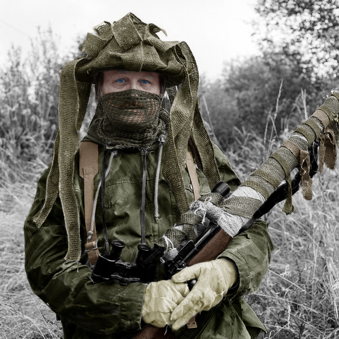 A person in camouflage clothing and gloves stands outdoors holding a wrapped rifle. Their face is partially covered by a netted mask, and they wear a helmet made from fabric strips. Bushes and tall grass are in the background.