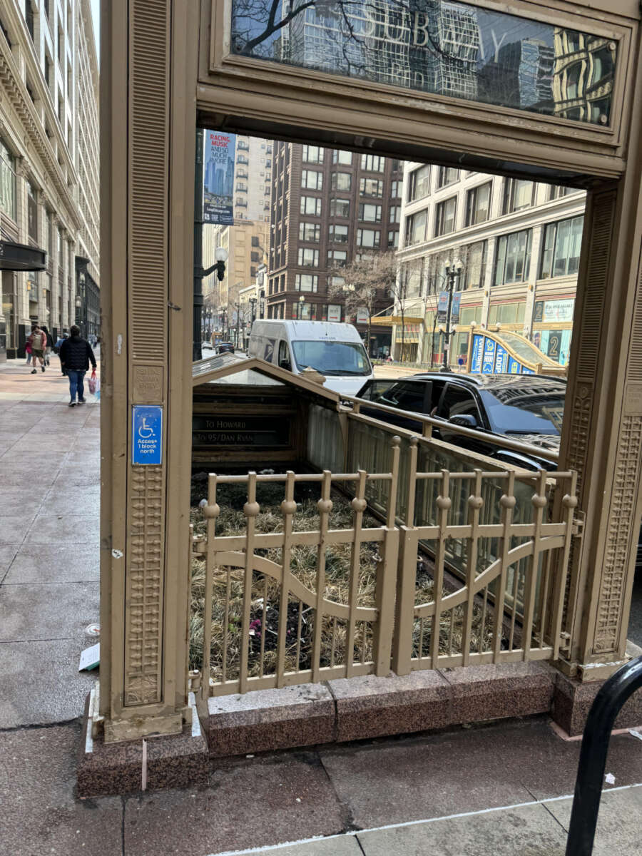 A Chicago subway entrance with gold fencing and a blue accessible entrance sign is on a busy city sidewalk surrounded by tall buildings, vehicles, and pedestrians on an overcast day.