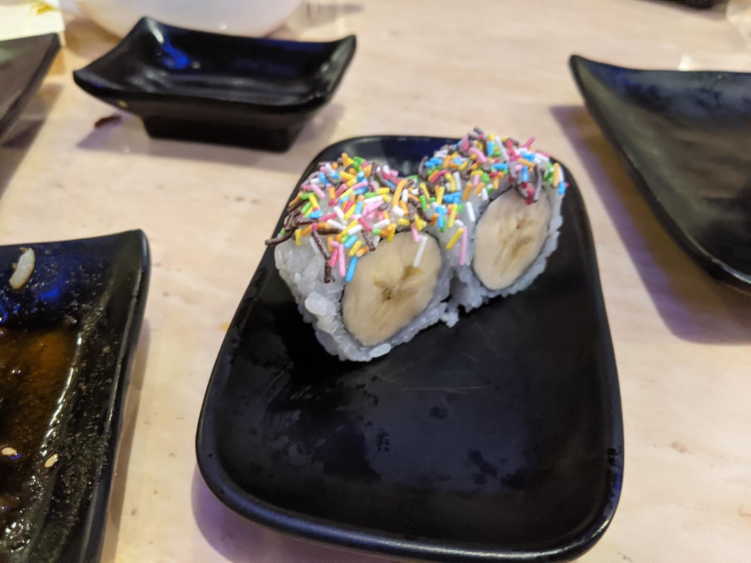 Two pieces of sushi filled with banana slices and topped with colorful sprinkles are served on a black plate. Other empty plates are visible in the background.