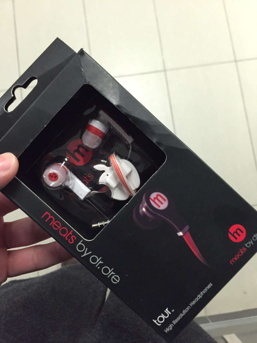 A hand holds a box of "meats by dr. dre" headphones, a parody of "beats by dr. dre." The headphones inside the box are white and red, shaped like pieces of meat. The background is a tiled floor.