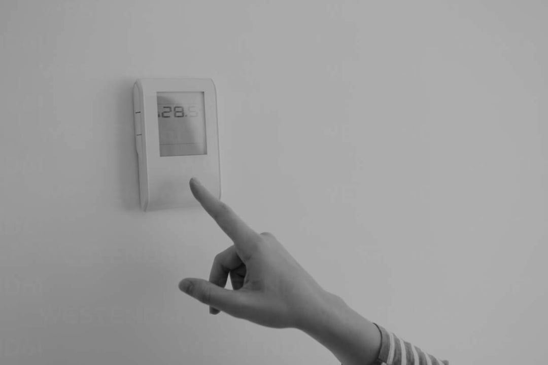 A person’s hand reaches toward a wall-mounted digital thermostat displaying a temperature of 28.5°C. The thermostat is attached to a plain, light-colored wall.