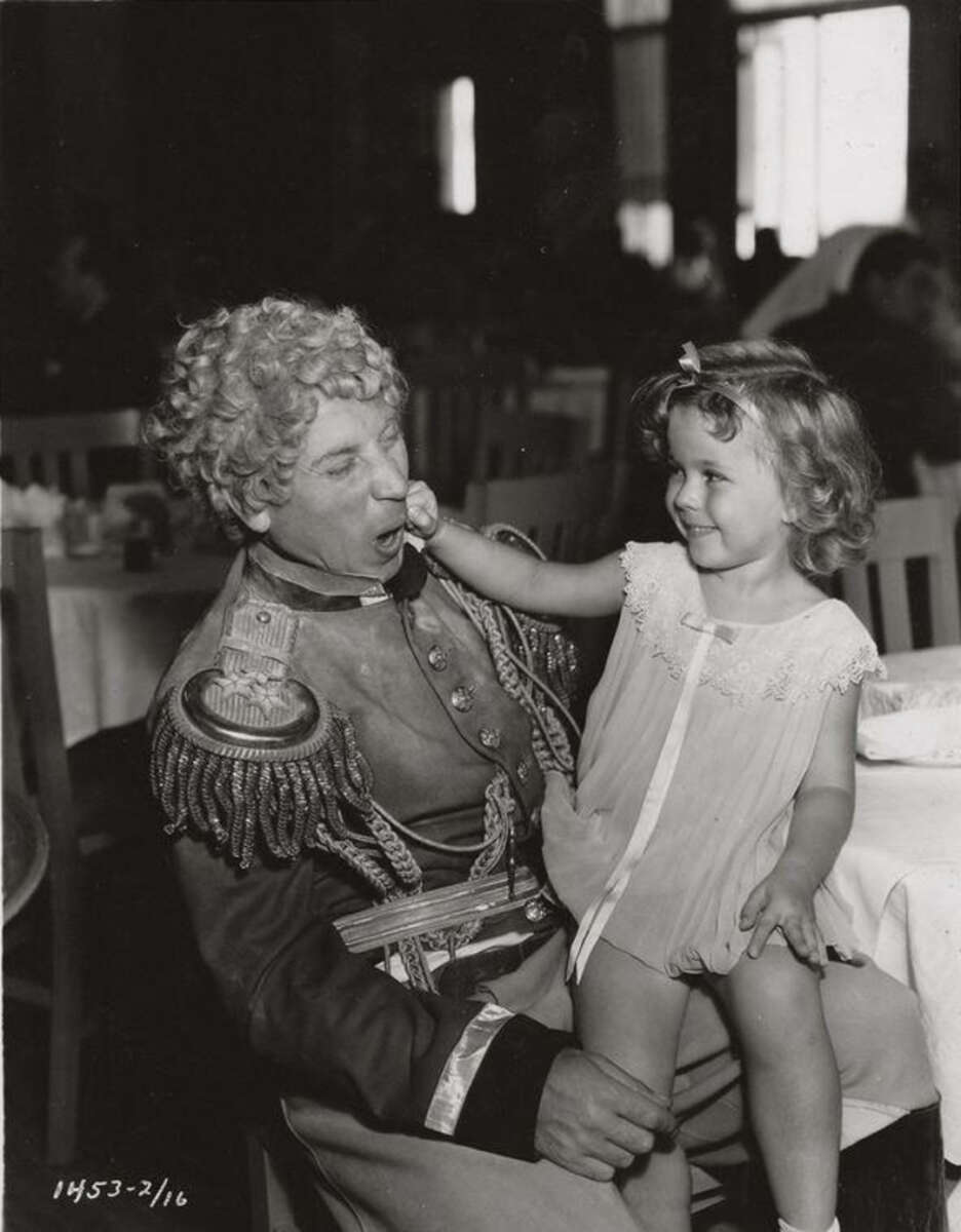 A man in a military costume with curly hair sits at a table, smiling as a young girl in a light dress playfully touches his nose. The two share a joyful moment in a lively, well-lit room.