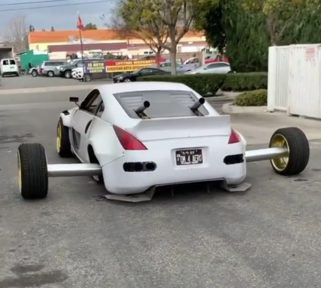 A modified white sports car with very wide rear wheels, yellow rims, and extended metal axles is parked in a parking lot. The car has a custom body kit and two rear exhaust pipes pointing upward.