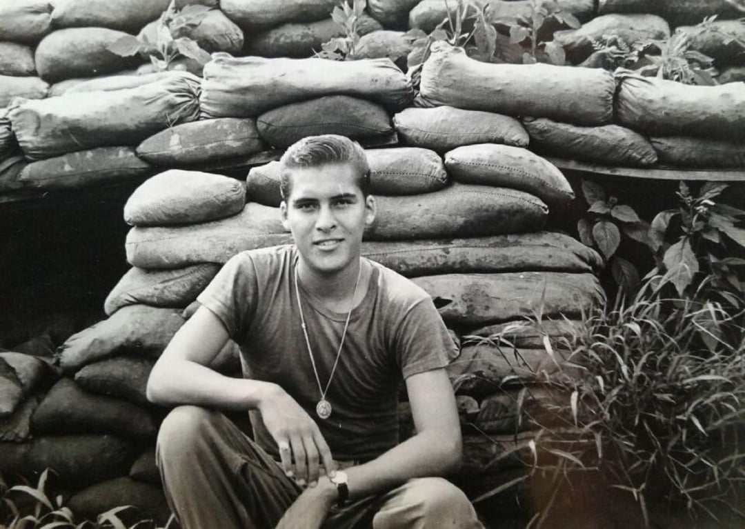 A young man in a T-shirt and military-style dog tags sits in front of a wall made of sandbags, surrounded by vegetation. The black and white photo has a vintage, historical feel.