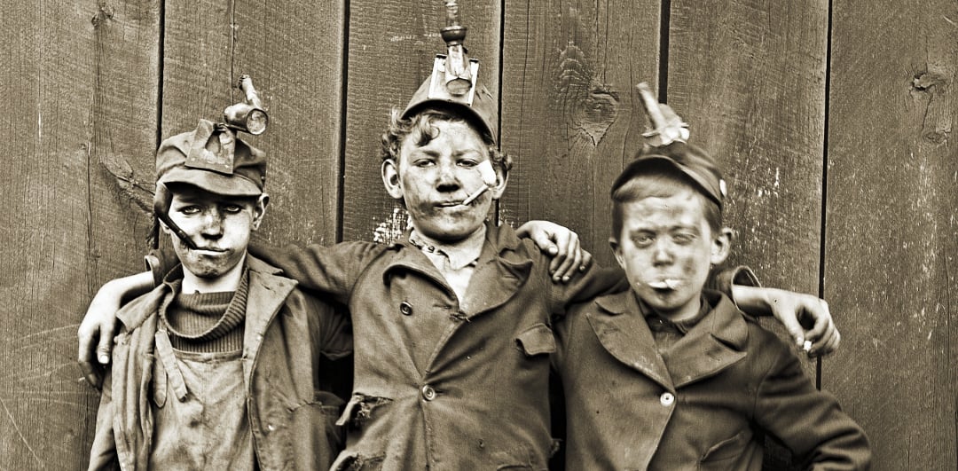 Three young boys in dirty work clothes and mining helmets stand close together, arms around each other, in front of a wooden wall. Their faces are smudged with dirt, and two have cigarettes in their mouths.
