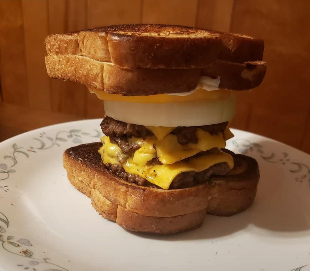 A stacked homemade sandwich on toasted bread with two beef patties, melted cheese, a thick slice of raw onion, and a fried egg, served on a white plate with a floral pattern.