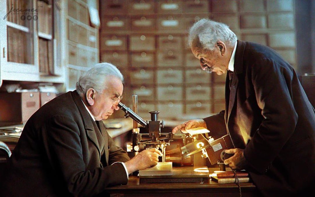 Two elderly men in suits work together in a vintage laboratory. One looks through a microscope while the other adjusts a light source on the table. Wooden drawers fill the background.