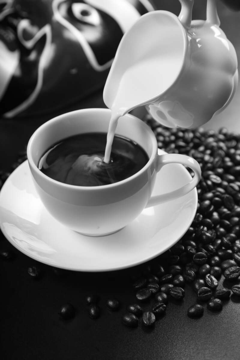 A black and white image of milk being poured from a small pitcher into a cup of coffee, with coffee beans scattered around the saucer and table.