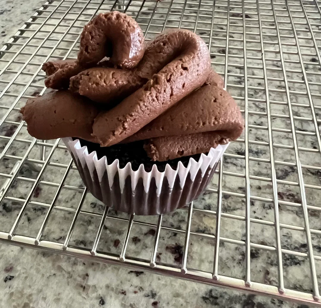 A chocolate cupcake in a white paper liner sits on a metal cooling rack, topped with a generous swirl of chocolate frosting. The background is a speckled granite countertop.