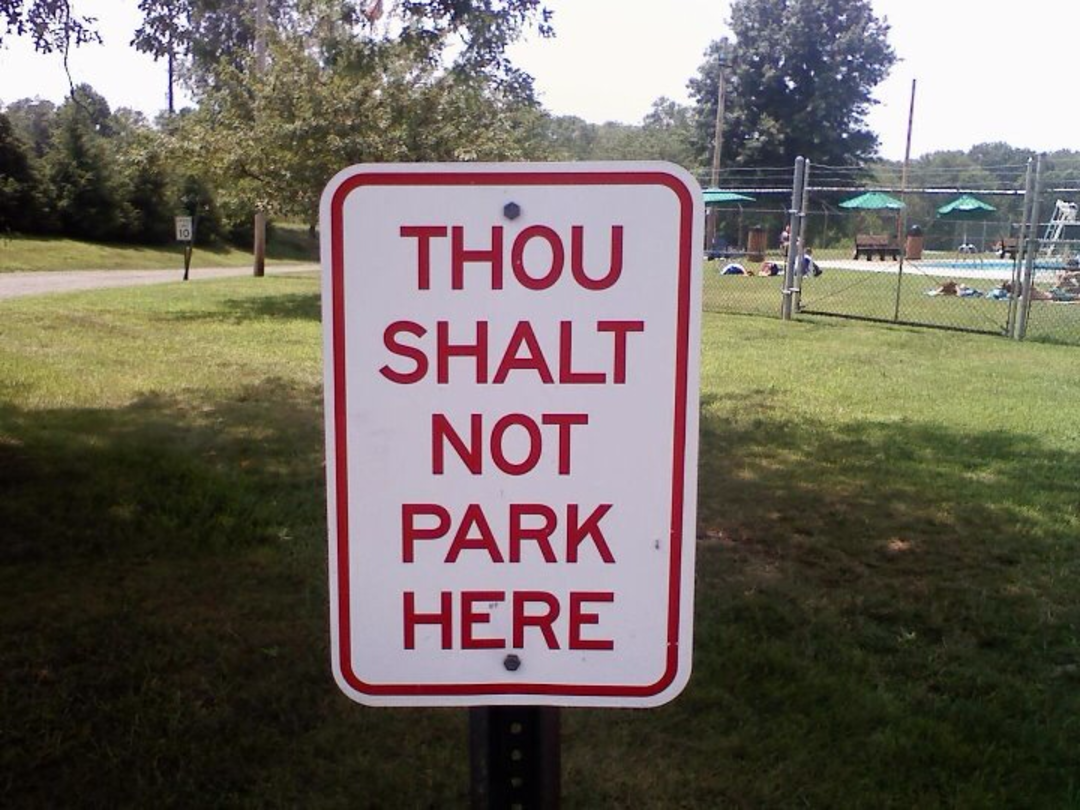 A white sign with red text reads "THOU SHALT NOT PARK HERE," placed on grass near a pool area with trees and a fence in the background.