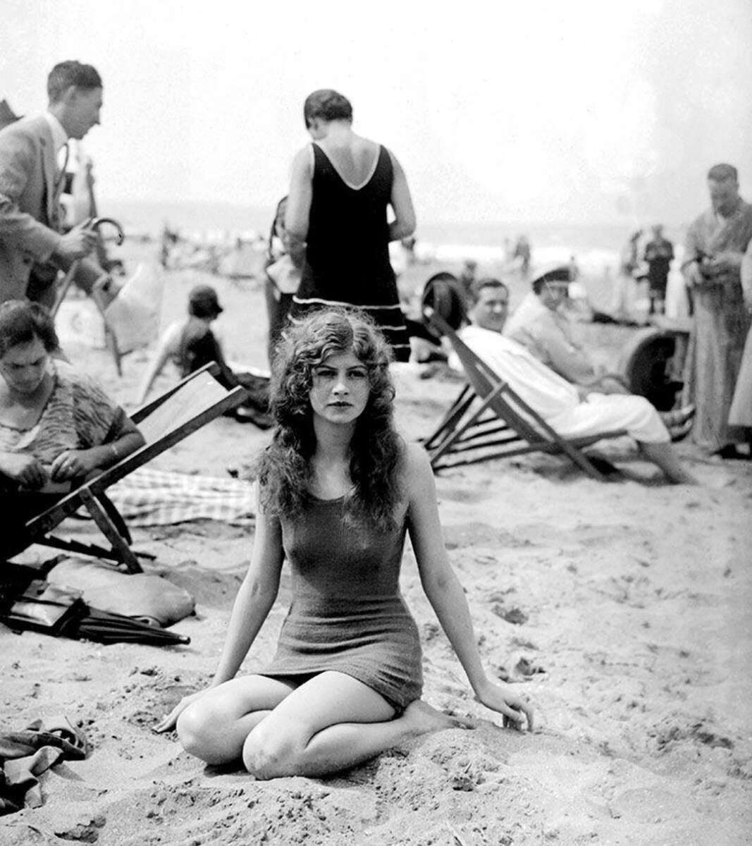 A young woman in a vintage swimsuit sits on sandy beach, looking at the camera. Several people in early 20th-century attire relax on deck chairs and walk in the background. The scene is lively and crowded.