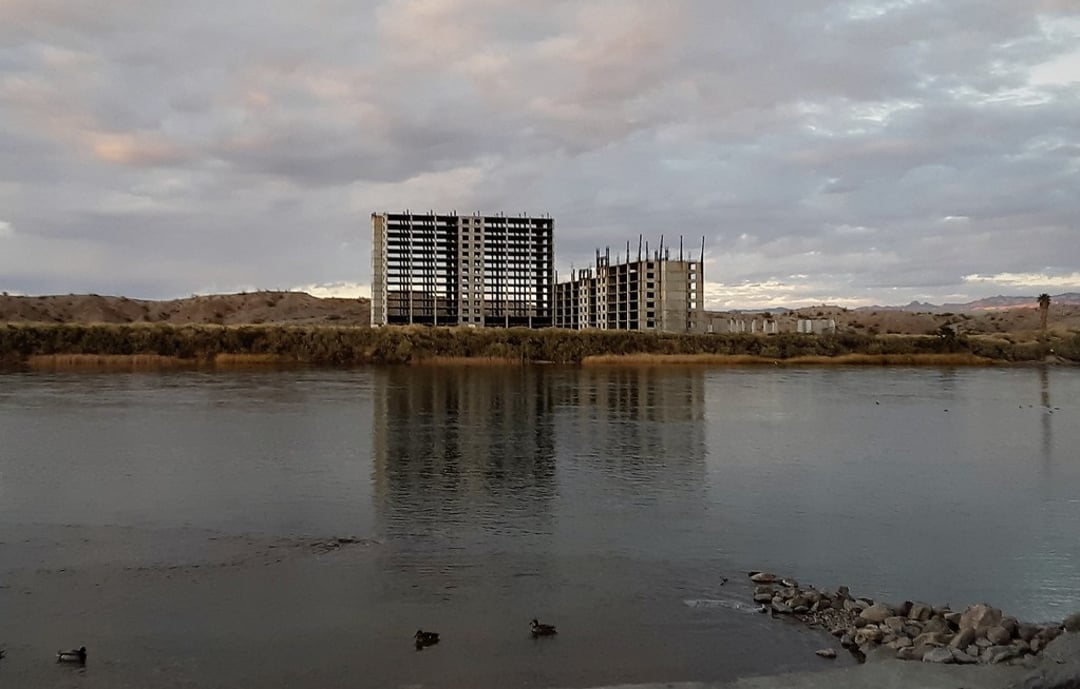 An unfinished, abandoned building stands near a calm river, with dry vegetation and distant hills in the background under a cloudy sky. Ducks are visible in the water in the foreground.