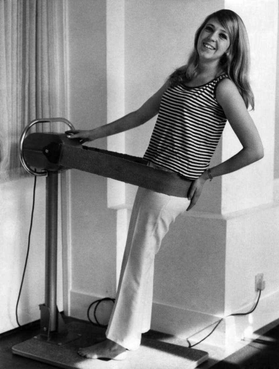 A woman in a striped sleeveless top and white pants stands barefoot, smiling while using a vintage vibrating belt exercise machine in a bright room with large windows.